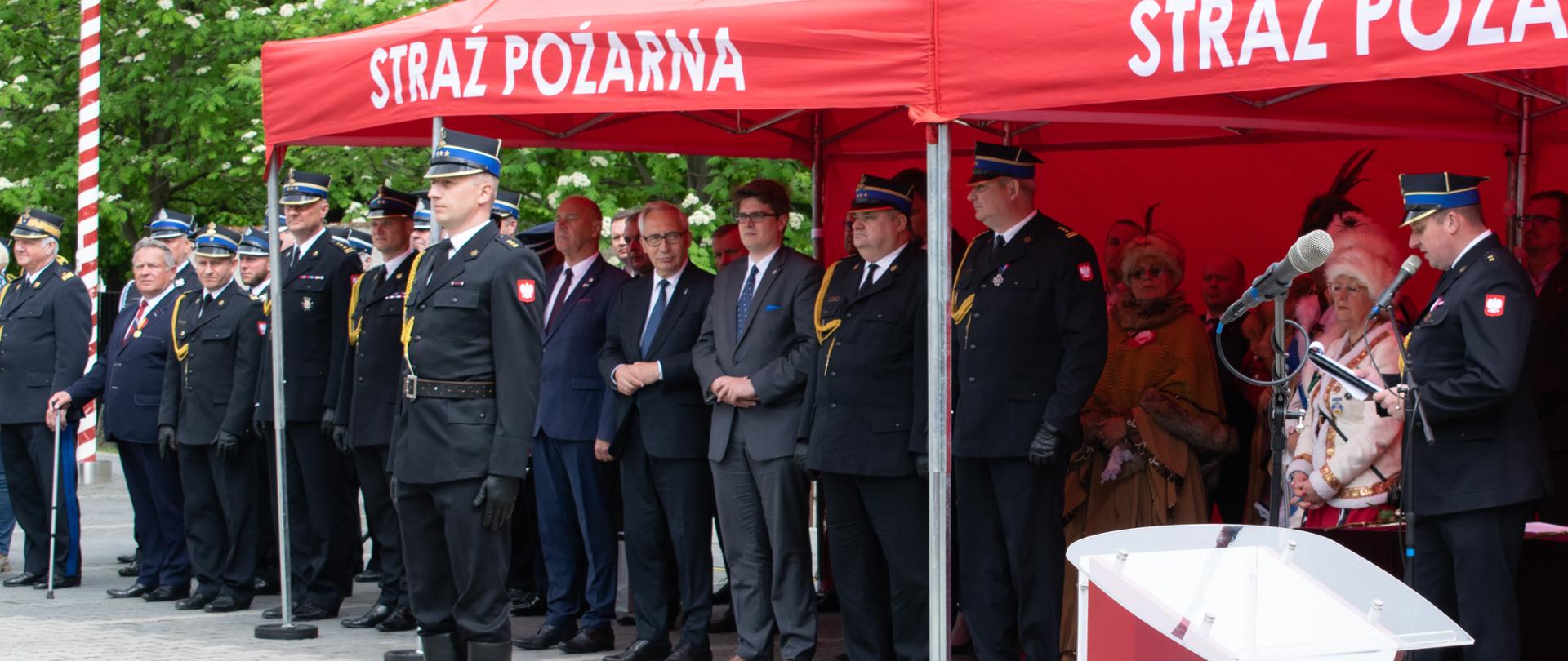 Widok na trybunę honorową gdzie stoją zaproszeni goście. Przed nimi dowódca uroczystości. Po prawej stronie narrator.