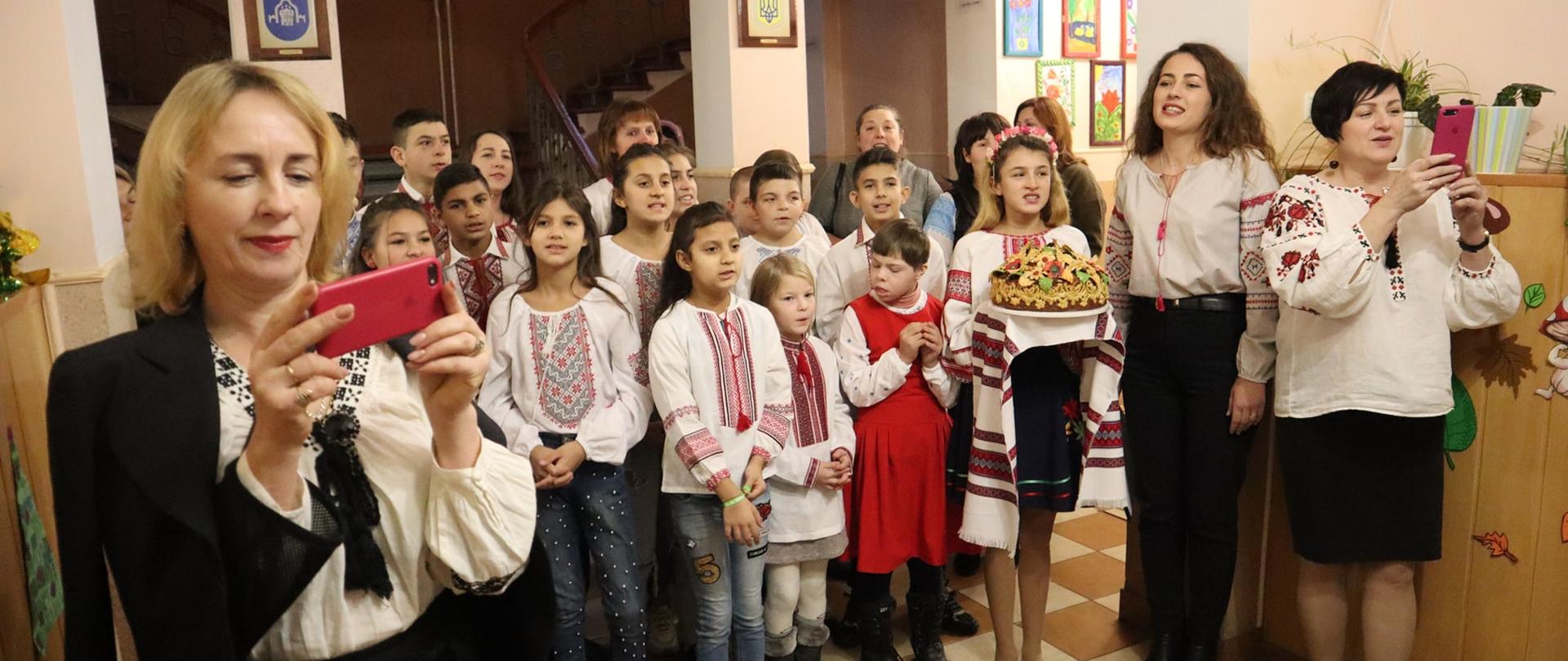Dzieci ubrane w tradycyjne stroje ukraińskie stoi w rzędzie. Jedna dziewczynka trzyma w rękach udekorowane ciasto. Trzy nauczycielki stoją obok grupy dzieci, robiąc zdjęcia komórką. 