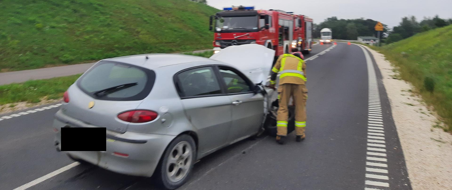 Zdjęcie przedstawia uszkodzony pojazd osobowy w obrębie, którego strażacy prowadzą działania. Obok pojazdu znajdują się wozy pożarnicze.. 