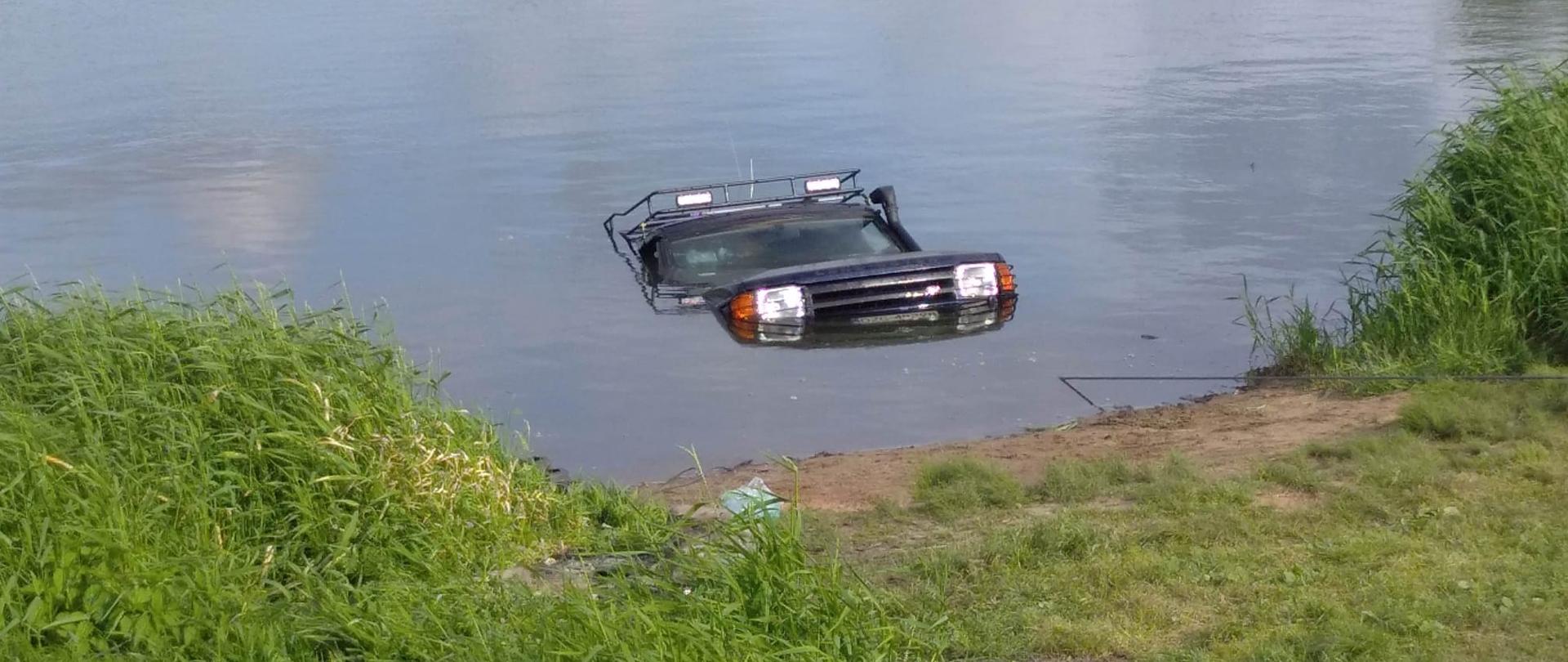 Dzień. Widok na brzeg akwenu. Brzeg częściowo porośnięty wysoką trawą. Wyraźne miejsce częściowo pozbawione roślinności ułatwiający dostęp do wody. Z wody wynurza się część samochodu terenowego - przednie światła, maska, szyba oraz część dachu.