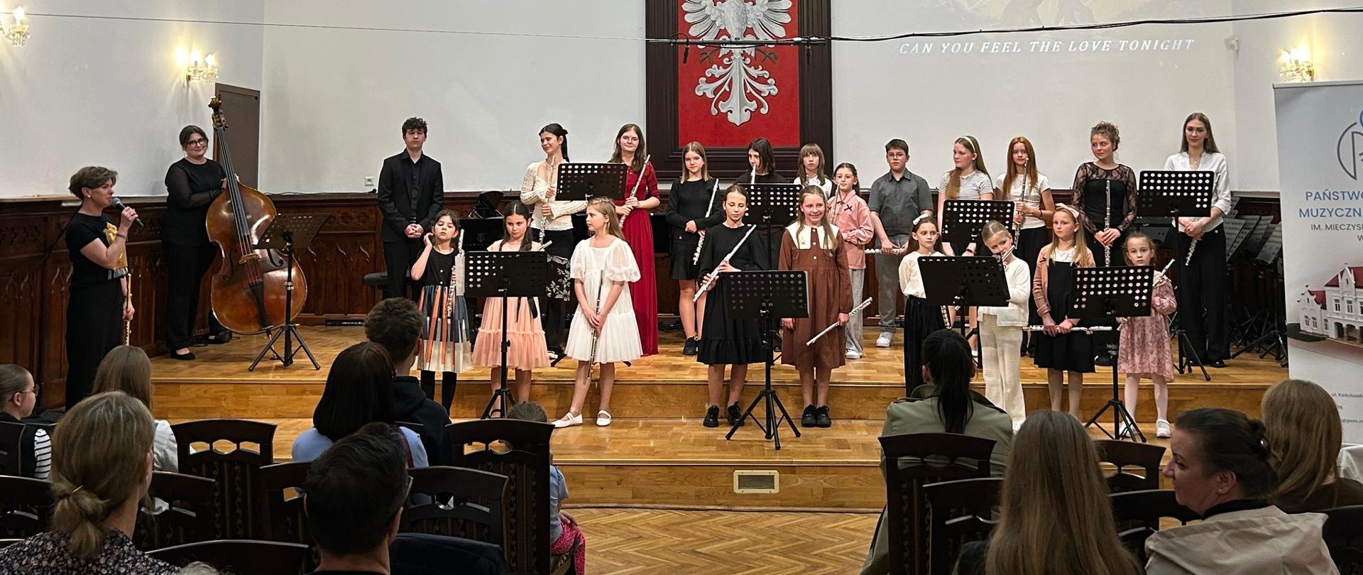 Grupa uczniów z fletami stoi na scenie Sali Królewskiej podczas koncertu muzyki fletowej, w tle herb Polski i portrety królów polskich.
