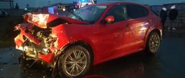 Zdjęcie przedstawia stojący na drodze samochód osobowy koloru czerwonego. Samochód ma uszkodzony przód. 