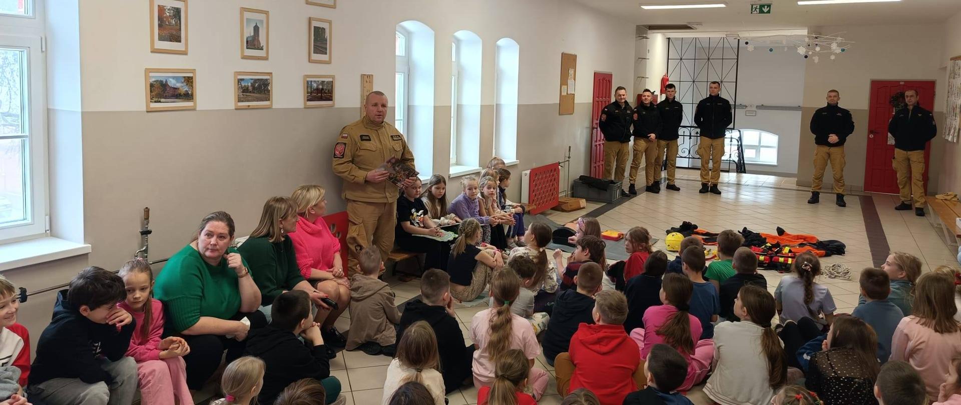 Szkoła Podstawowa nr 5 im. Ziemi Braniewskiej w Braniewie,korytarz szkolny, dzieci siedzą na i słuchają strażaka. Dalej w oddali kolejni strażacy.