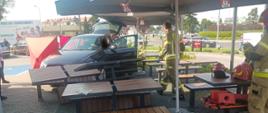 Widok z przodu. Samochód osobowy w ogródku restauracyjnym. Na pierwszym planie przewrócone stoły. Dalej samochód z otwartymi drzwiami, w środku siedzi kierowca. Strażacy podchodzą do poszkodowanej celem udzielenia pomocy. Za samochodem rozłożony parawan. 