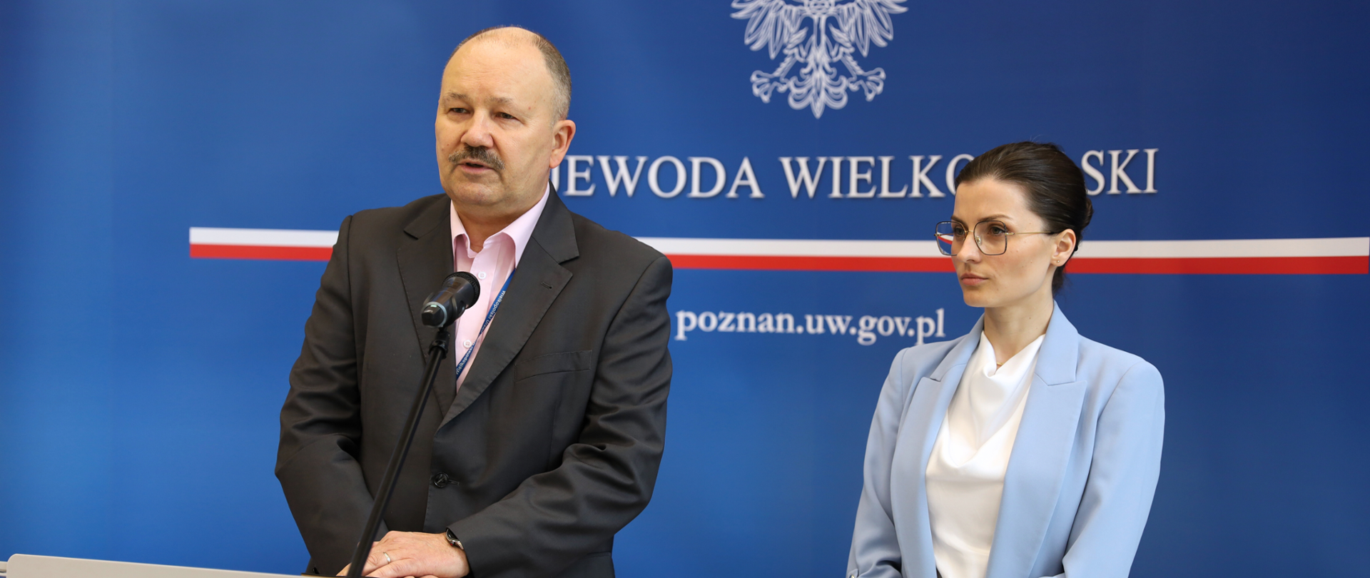 Mężczyzna stoi przy mównicy z orłem i przemawia. Obok niego kobieta ubrana w białą bluzkę i błękitny żakiet