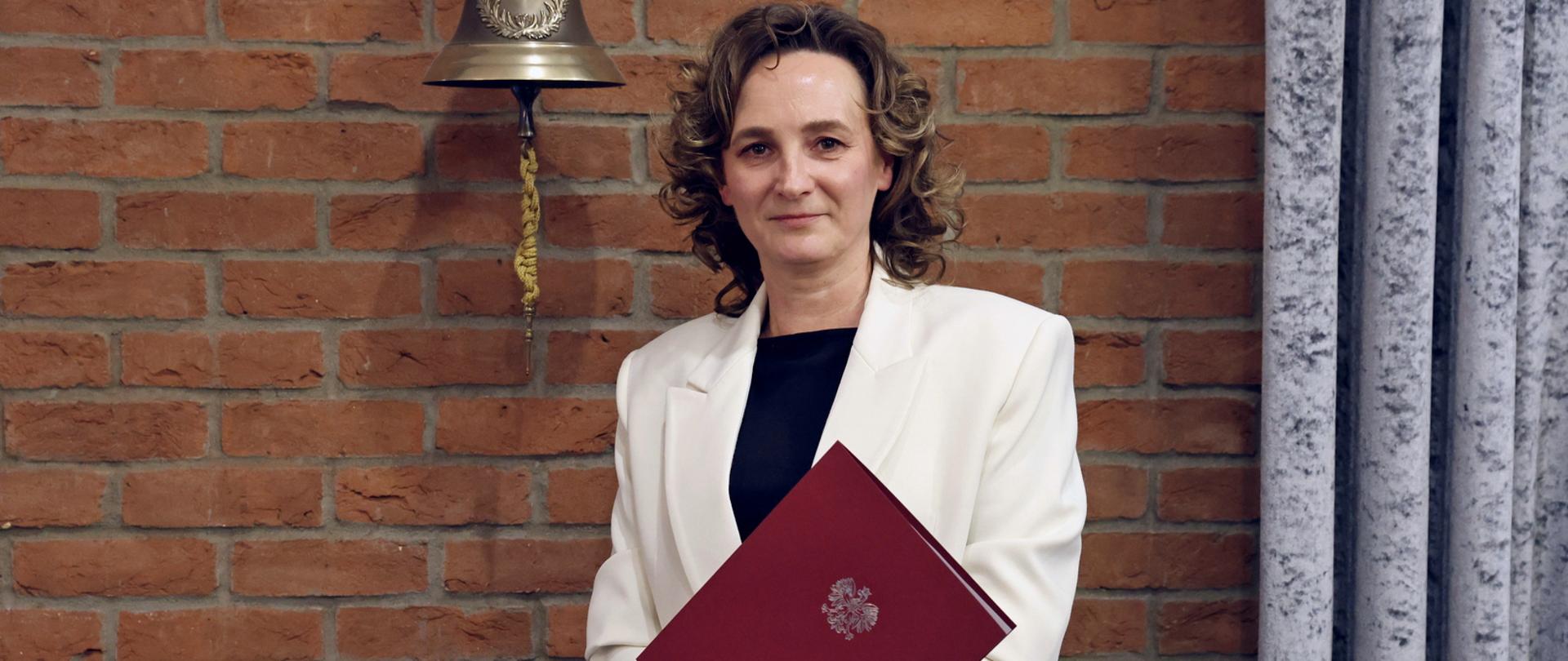 Gabinet Komendanta Szkoły, kobieta trzymająca czerwoną teczkę stojąca na tle ceglanej ściany