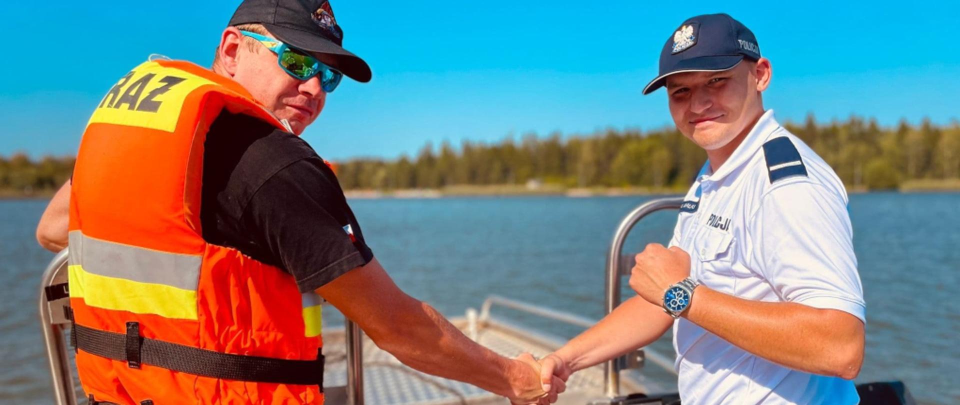 Zdjęcie przedstawia funkcjonariusza KM PSP w Jaworznie oraz funkcjonariusza KMP w Jaworznie podających sobie prawe dłonie na łodzi motorowej w czasie patrolowania zalewu Sosina.