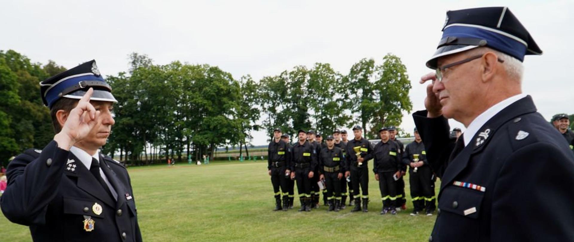 Stadion sportowy w Kluczewsku, komendant gminny OSP dh. Dariusz Ławiński składa meldunek o zakończeniu zawodów Wójtowi Gminy Kluczewsko dh Rafałowi Pałce, w tle drużyny OSP uczestnicy zawodów