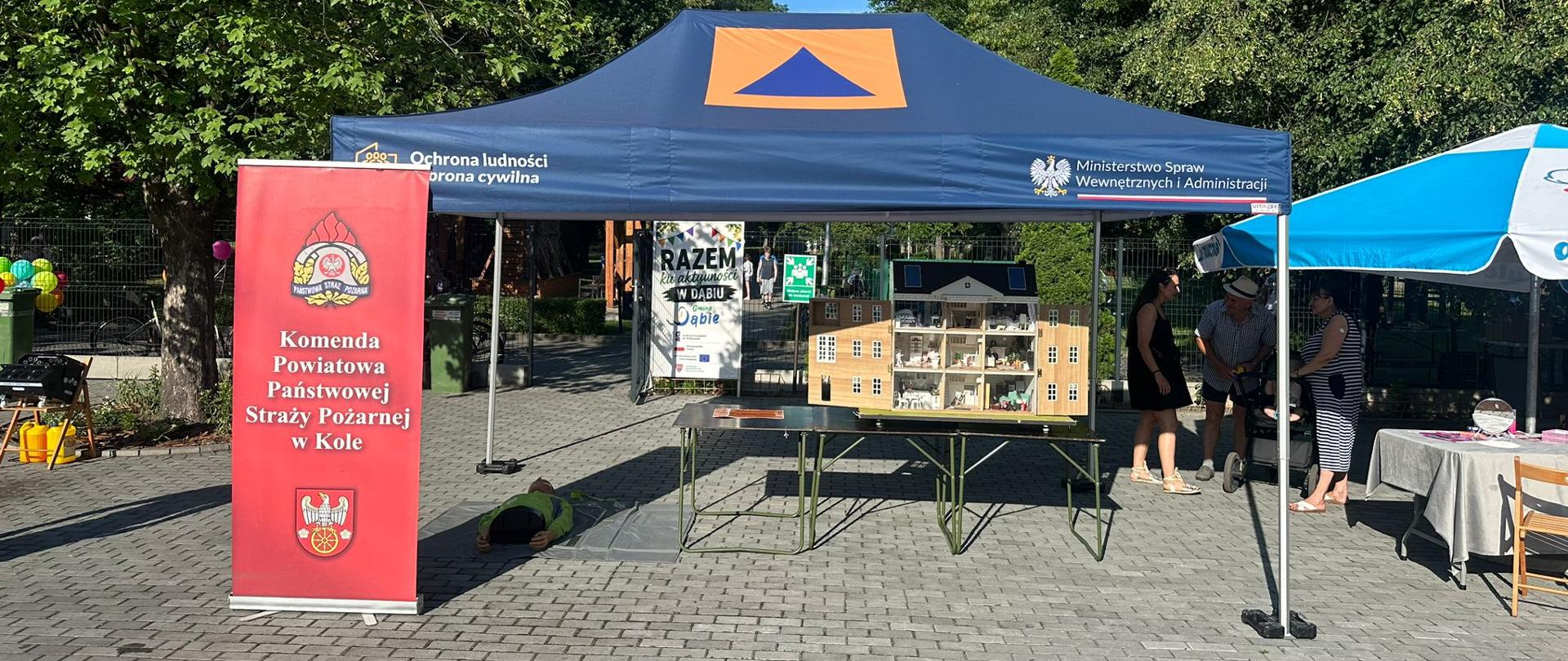Stoisko edukacyjne Komendy Powiatowej Państwowej Straży Pożarnej w Kole podczas festynu plenerowego. Pod granatowym namiotem oznaczonym symbolami obrony cywilnej i Ministerstwa Spraw Wewnętrznych i Administracji znajduje się makieta domu służąca do prezentacji zagrożeń pożarowych. Obok stoi czerwony baner PSP, a wokół widoczne są inne stoiska i uczestnicy wydarzenia.