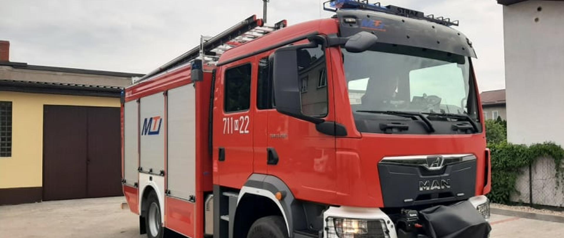 Czerwony samochód ratowniczo-gaśniczy stoi na placu wewnętrznym komendy. W tle budynek garażowy.