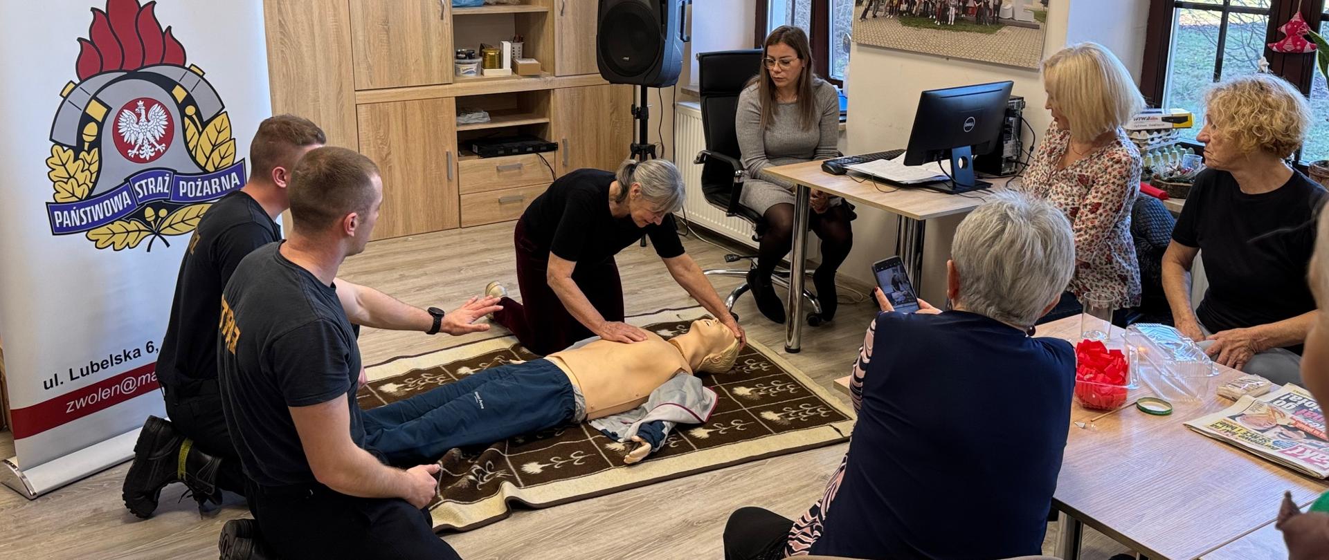 Zdjęcie przedstawia szkolenie z pierwszej pomocy, prawdopodobnie dla dorosłych, odbywające się w pomieszczeniu Komendy Powiatowej Państwowej Straży Pożarnej w Zwoleniu. Na pierwszym planie widać dwie osoby, klęczącą obok manekina do resuscytacji i demonstrującą techniki pierwszej pomocy. Obok nich klęczy dwóch strażaków obserwujących pokaz. Na drugim planie siedzi grupa osób, w tym kilka kobiet, które obserwują szkolenie. W tle widoczny jest baner z logo i danymi kontaktowymi Komendy Powiatowej Państwowej Straży Pożarnej w Zwoleniu.