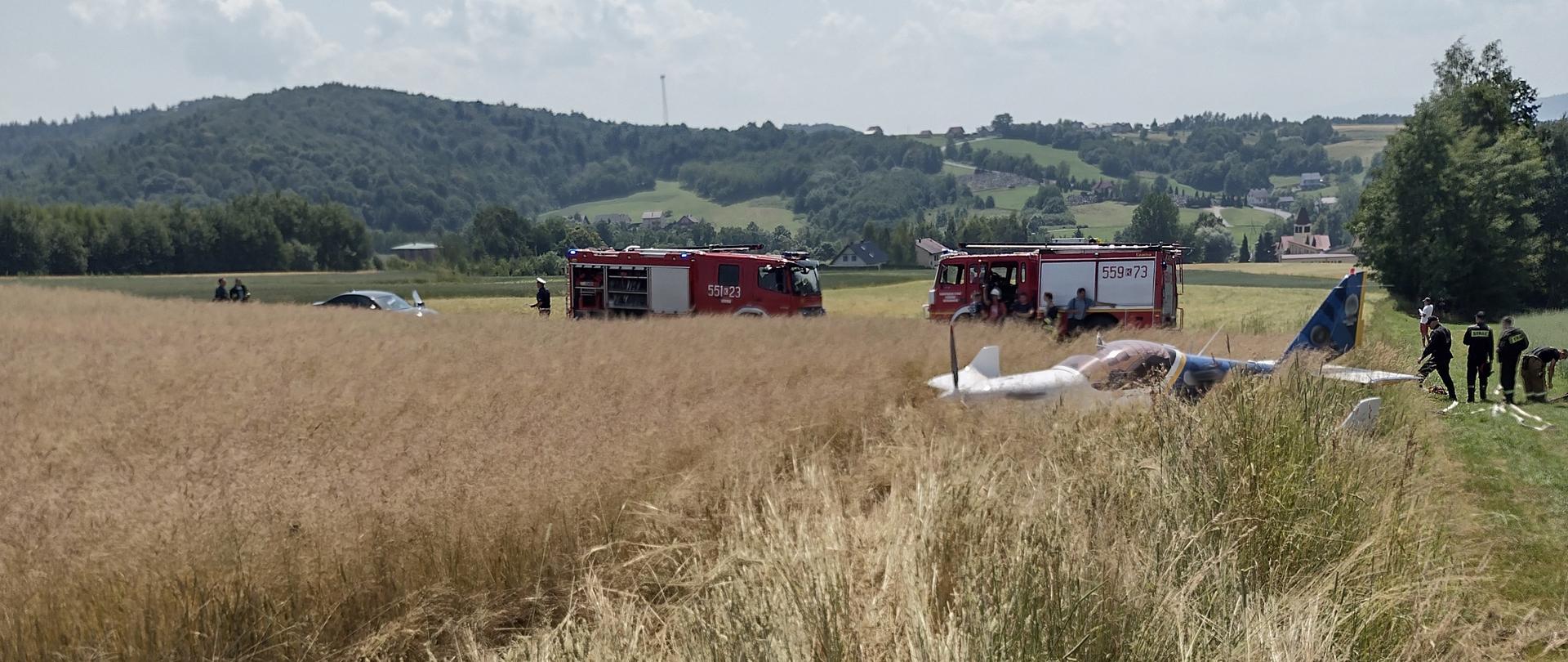 Pole uprawne (zboże). Po prawej stronie grupa strażaków i widoczne węże pożarnicze. W zbożu awionetka (jednosilnikowy dwumiejscowy dolnopłat). W głębi dwa samochody ratowniczo- gaśnicze. W tle widok na miejscowość i pagórkowaty teren.