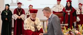 Minister Wieczorek stoi naprzeciwko mężczyzny w czerwonym ceremonialnym stroju z gronostajowym kołnierzem, podaje nu rękę, dalej stoi szereg osób w ceremonialnych strojach, obok bukiety kolorowych kwiatów.