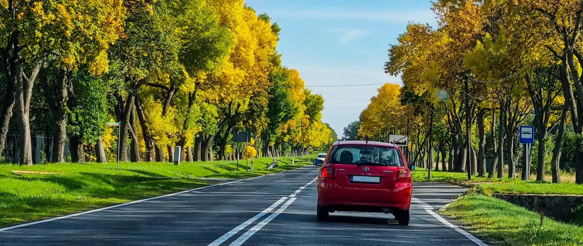Ciepły jesienny dzień. Czerwony samochód jadący drogą. Po obu stronach drogi drzewa, po lewej zatoka autobusowa.