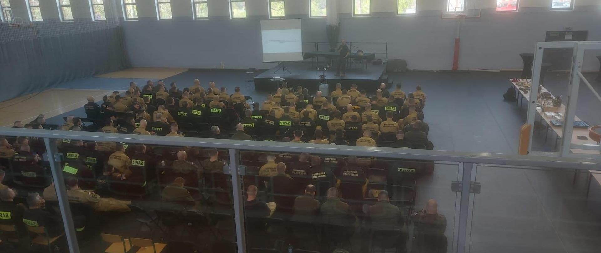 Zdjęcie przedstawia strażaków oraz wykładowcę podczas wykładu na sali gimnastycznej, w tle widać uruchomioną na projektorze prezentację
