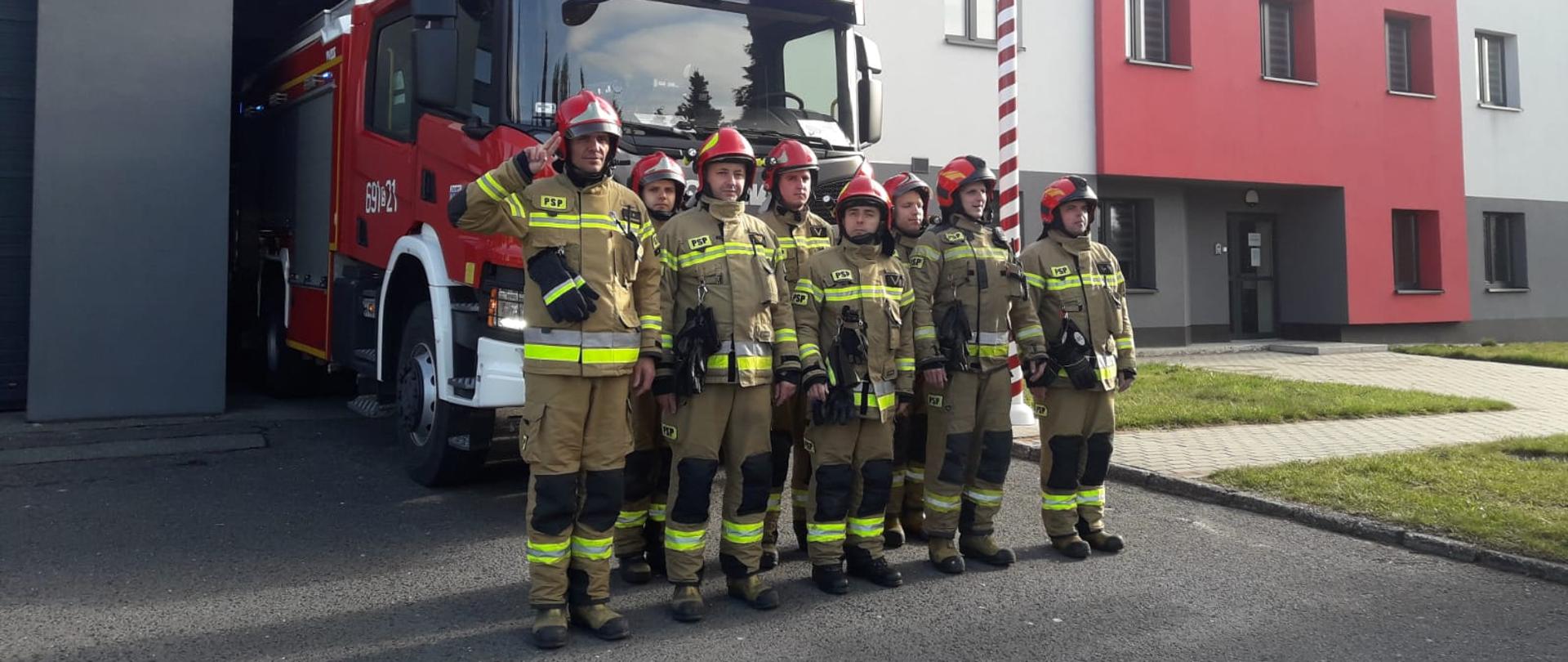 Strażacy w mundurach bojowych i hełmach stoją w dwuszeregu na tle samochodu strażackiego i remizy. Pierwszy z nich salutuje. 