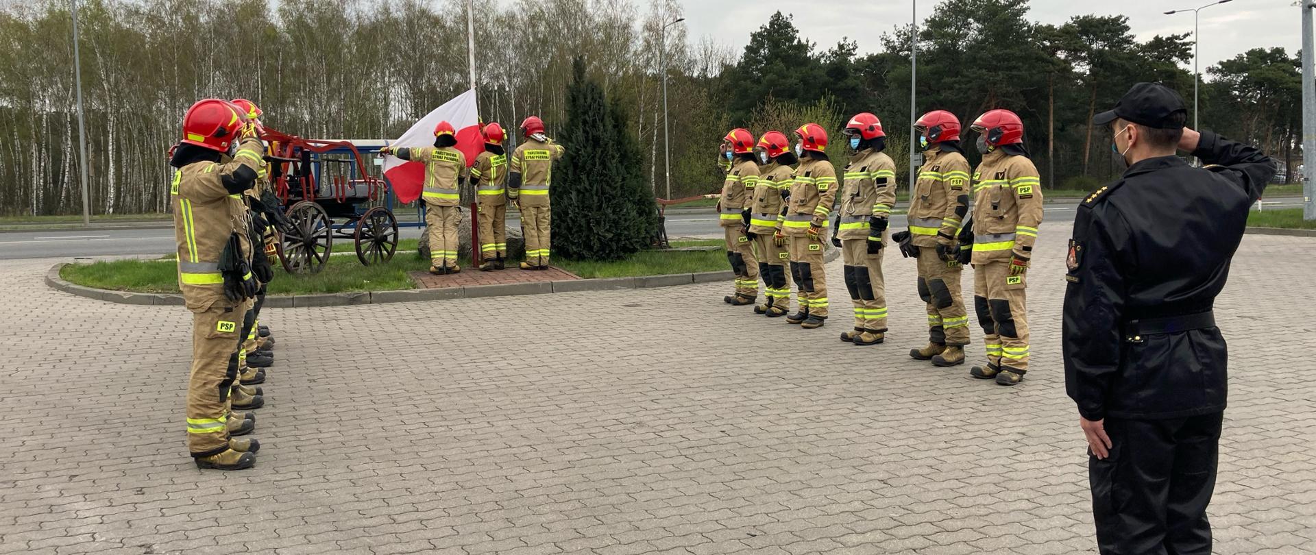 Strażacy w ubraniach specjalnych stojący przy maszcie, na którym jest zawieszona flaga Polski, wszyscy maseczki na twarzach. 