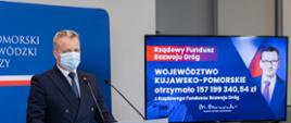 Wojewoda przy mównicy, w tle ekran, na którym napisane jest: Rządowy Fundusz Rozwoju Dróg, Województwo Kujawsko-Pomorskie otrzymało 157 199 340, 54 zł 