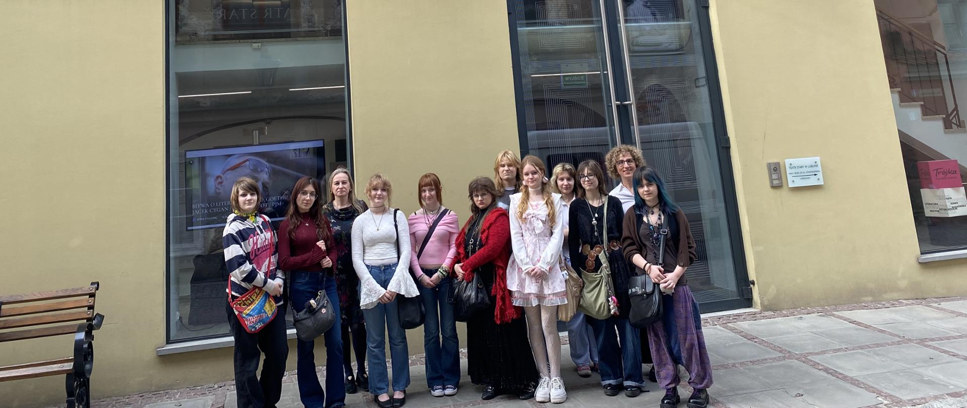 Grupa dziesięciu dziewcząt z dwiema wychowawczyniami pozuje do zdjęcia na tle fasady budynku teatru. Za nimi widać dwa duże okna i przeszklone drzwi, pomiędzy nimi wiszą lampy. Na górze na środku jest napis TEATR STARY, a po bokach są płaskorzeźby w białym kamieniu przedstawiające dwa psy ze skrzydłami po bokach kielicha z owocami, a nad nim głowa.