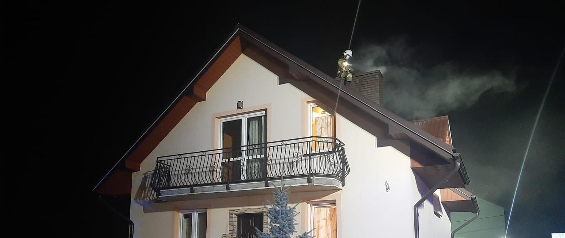 Budynek mieszkalny z komina wydobywa się dym a obok komina na dachu strażak prowadzący swoje działania. 
