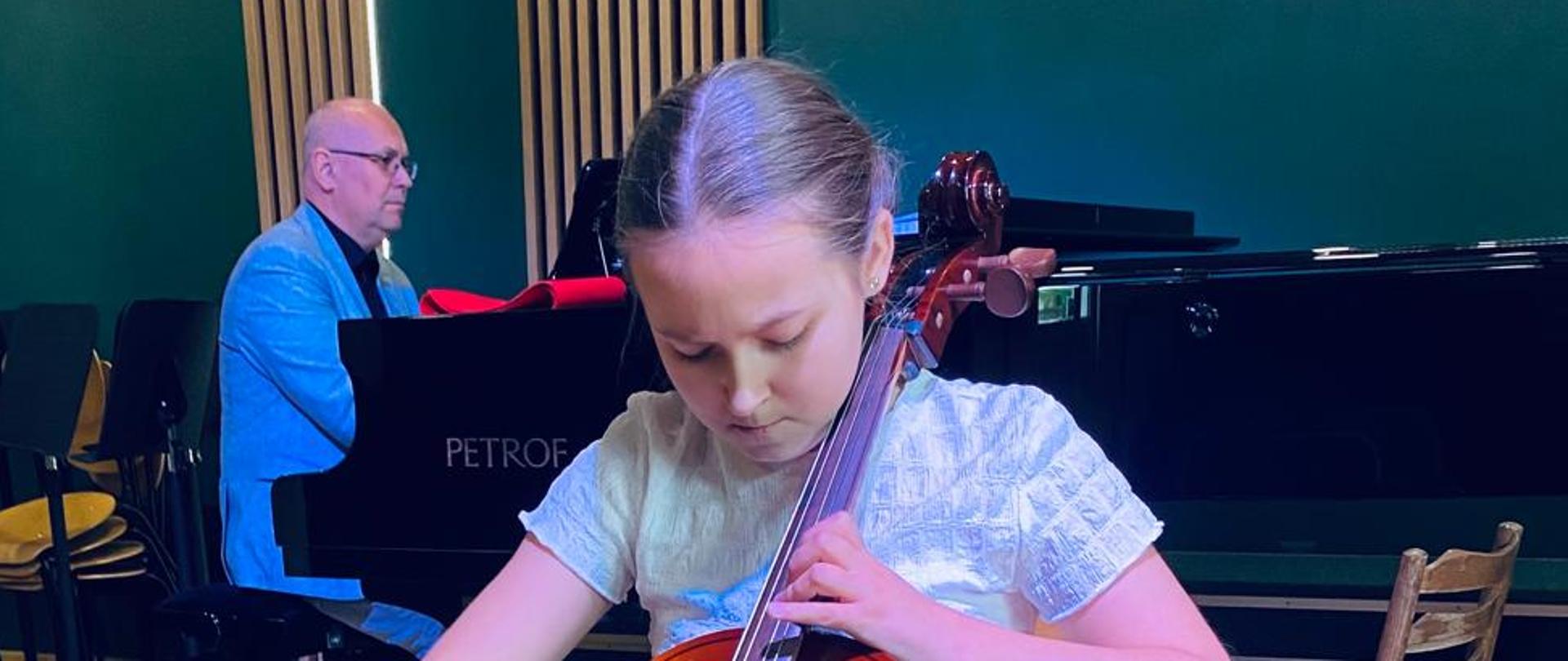 Na zdjęciu widać uczennicę która gra na wiolonczeli. Za nią stoi fortepian przy którym siedzi akompaniator. W tle widać zieloną ścianę z drewnianymi lamelami.