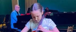 Na zdjęciu widać dziewczynkę która gra na wiolonczeli a w tle widoczny jest pianista który gra na fortepianie za nim widoczna jest zielona ściana.