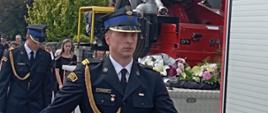 Samochód pożarniczy z trumną zmarłego jedzie w kondukcie pogrzebowym. Obok idą strażacy. w oddal drzewa i dach domu mieszkalnego. 