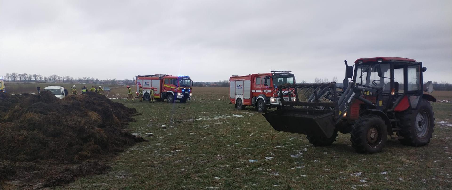 Na polu stoją dwa duże samochody strażackie, z prawej strony ciągnik rolniczy. Pochmurnie.