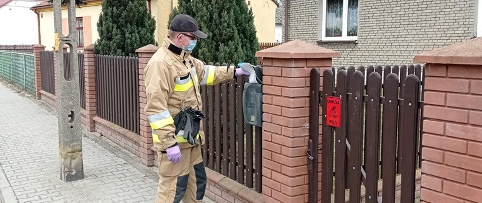 Strażak wrzuca ulotkę do skrzynki na listy znajdującej się na ogrodzeniu posesji 