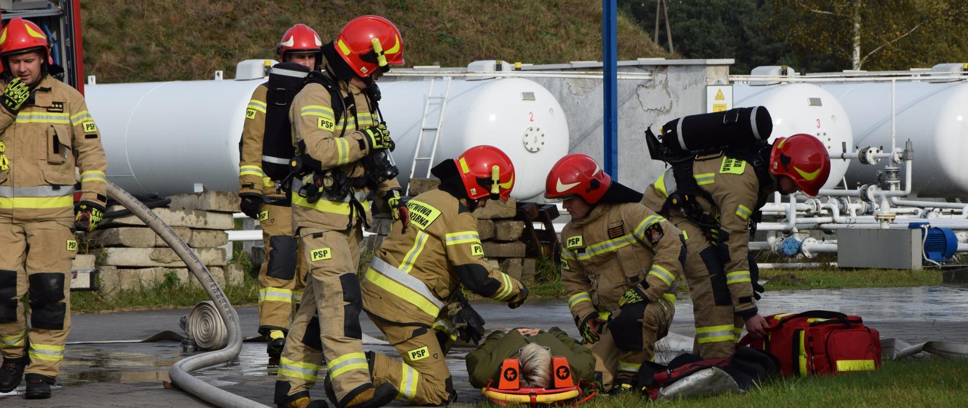 Na zdjęciu widać 6 strażaków ubranych w ubrania specjalne. Jeden z nich rozmawia przez radiostację nasobną, dwóch klęczy nad osobą poszkodowaną leżącą na desce ortopedycznej, kolejny schyla się do torby medycznej, następni dwaj stoją obok. W tle zbiorniki z gazem.