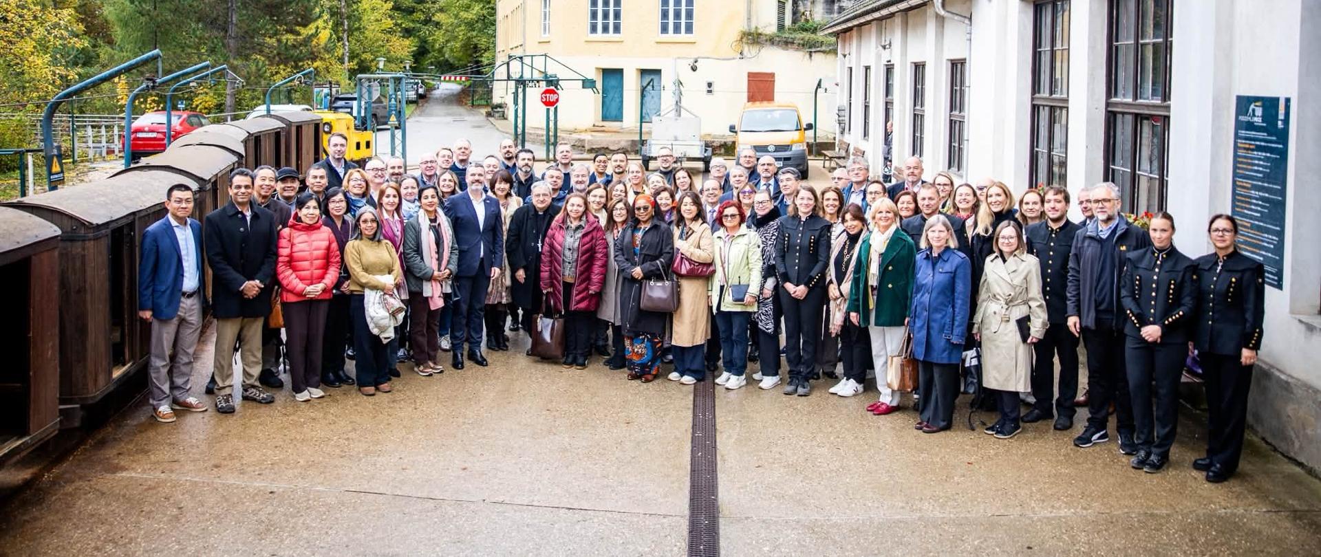 Duża grupa kilkudziesięciu osób pozuje do wspólnego zdjęcia na zewnątrz, przed budynkami dawnej kopalni w Mežicy. Po lewej stronie stoją niewielkie wagoniki górnicze na torach, po prawej widać biały budynek z dużymi oknami. W tle znajdują się zabudowania i zalesione wzgórza w jesiennych barwach.