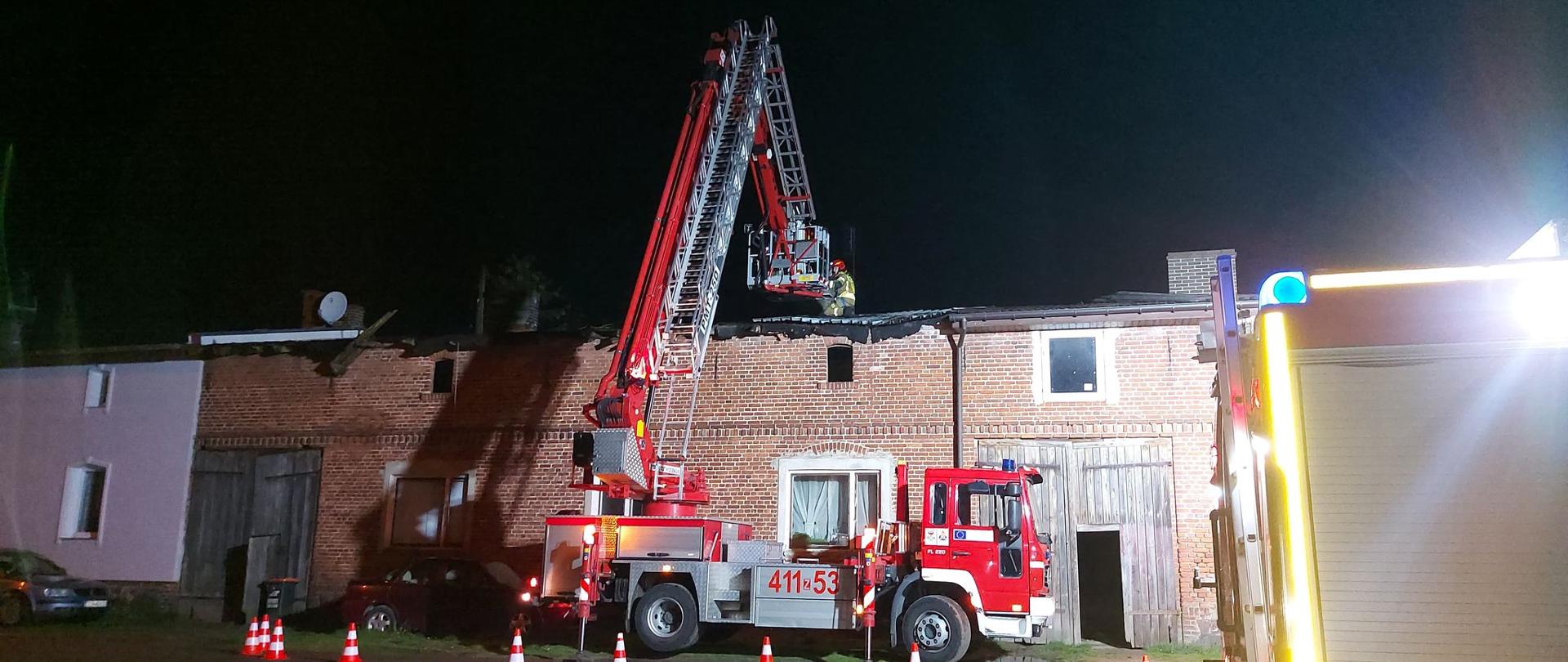 Na fotografii znajduje się rozstawiony podnośnik strażacki, który stoi obok budynku. Na dachu budynku pracują strażacy. Teren dookoła jest oświetlony, ponieważ działania trwają w porze nocnej.