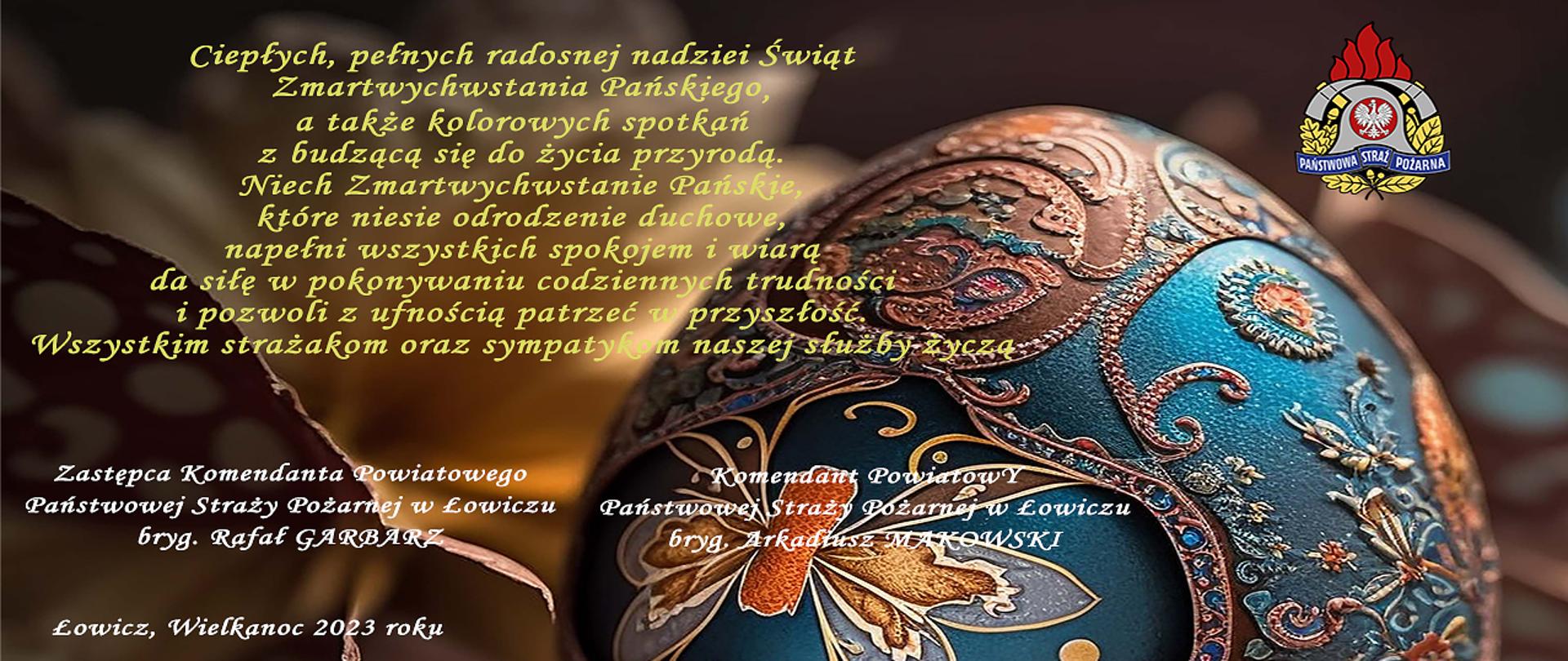 Pisanka koloru brązowego i zielononiebieskiego ozdobnie inkrustowana malowana ręcznie w motywy kwiatów u góry po prawej stronie logo państwowej straży pożarnej po stronie lewej życzenia świąteczne podpisane przez zastępcę komendanta powiatowego psp w łowiczu oraz przez komendanta powiatowego psp w łowiczu