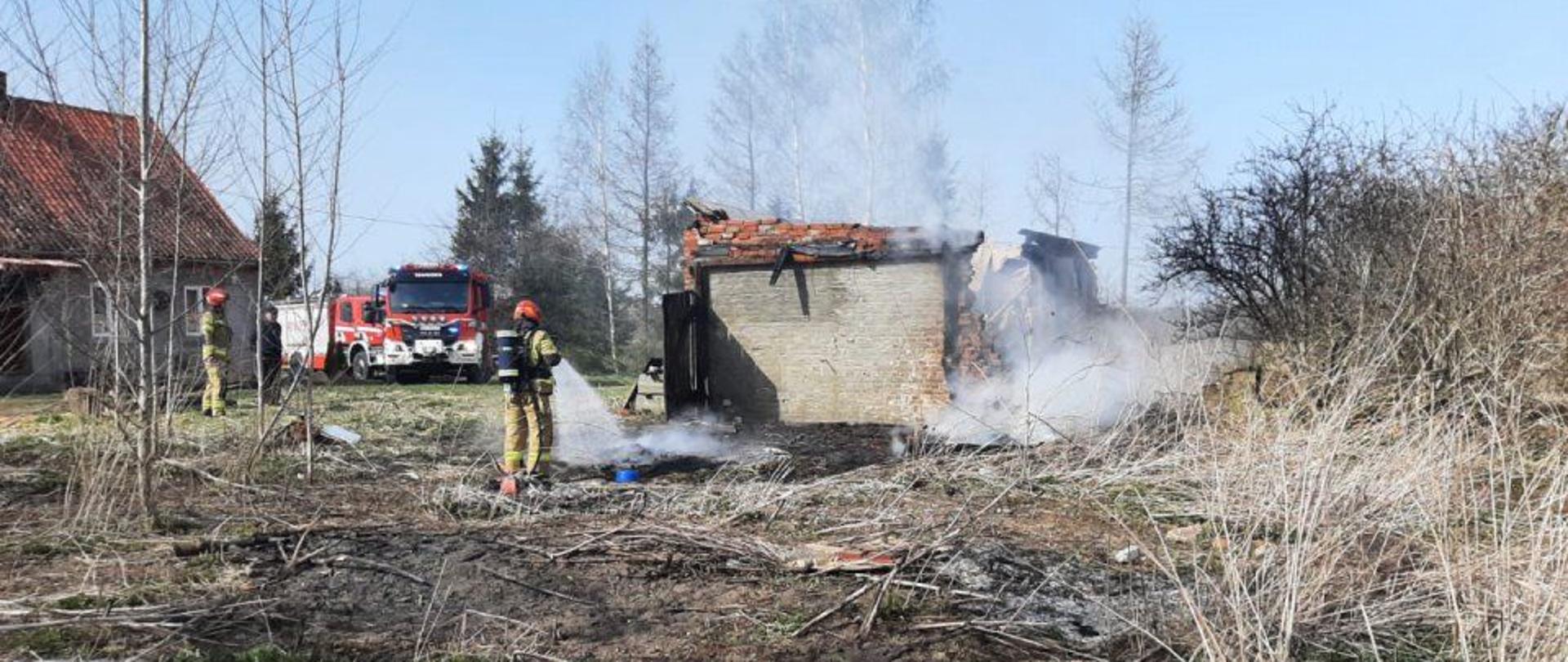 Zniszczony budynek gospodarczy po pożarze, dym. W oddali samochody strażackie