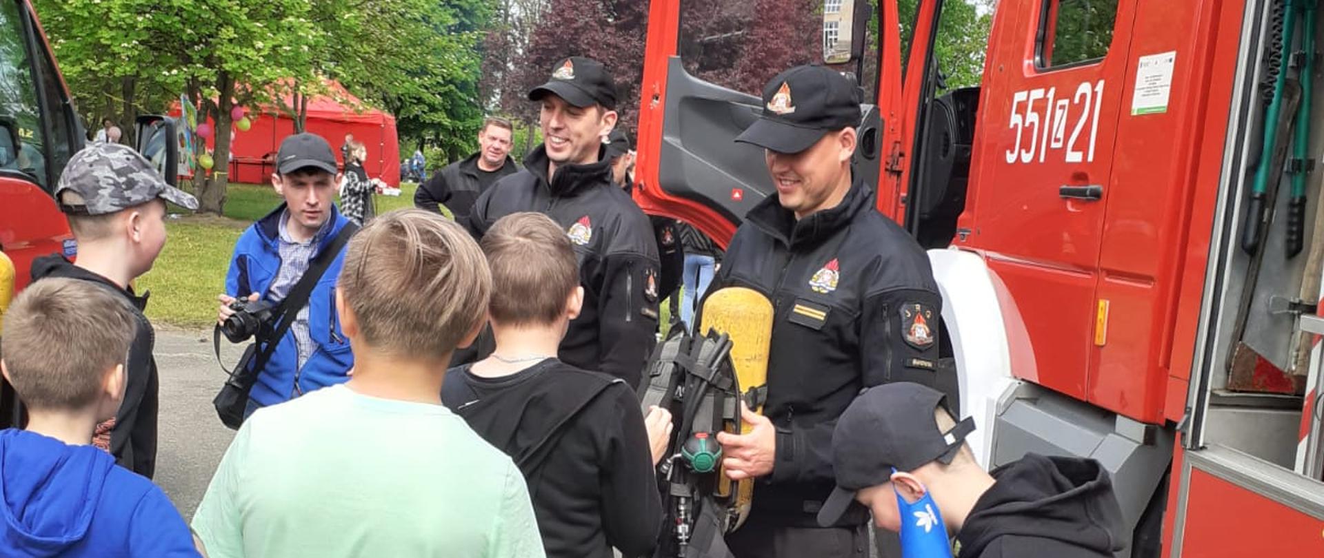Zdjęcie przedstawia pokaz sprzętu przez strażaków z okazji Międzynarodowego Dnia Dziecka. Strażacy pokazują butle powietrzne. Przed nimi znajduję się kilku zaciekawionych chłopców. Po bokach widzimy czerwone wozy strażacki.