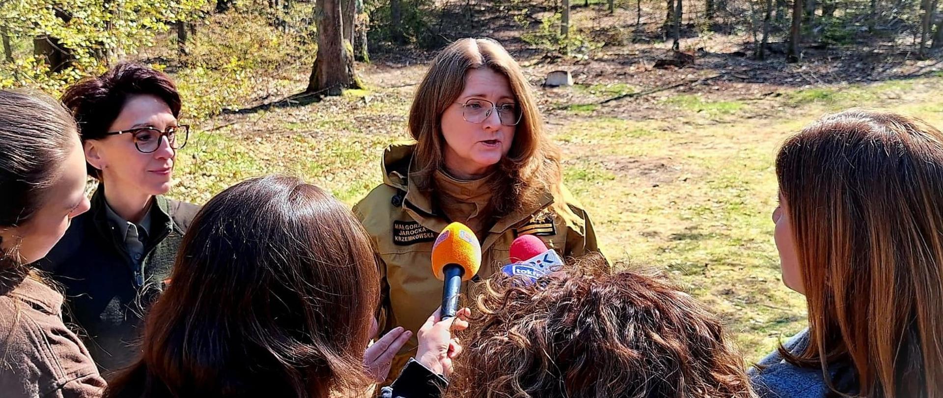 Kobieta strażak, a przed nią kilka osób z mikrofonami, skierowanymi w jej stronę. W tle las.