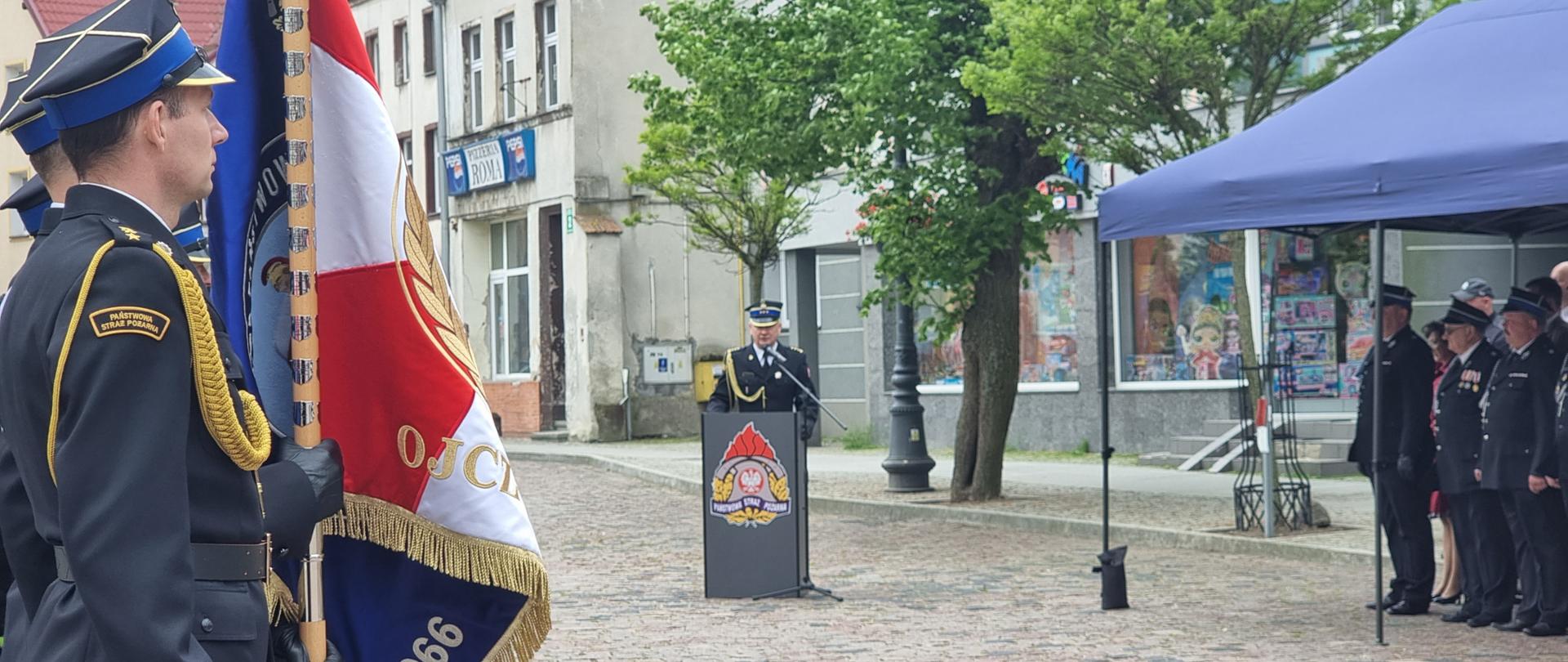 Pora dzienna, na placu po lewej stronie widać poczet sztandarowy, na środku stoi mównica z przemawiającym zastępcą komendanta wojewódzkiego. Po prawej stronie widać namiot pod którym stoją zaproszeni goście 