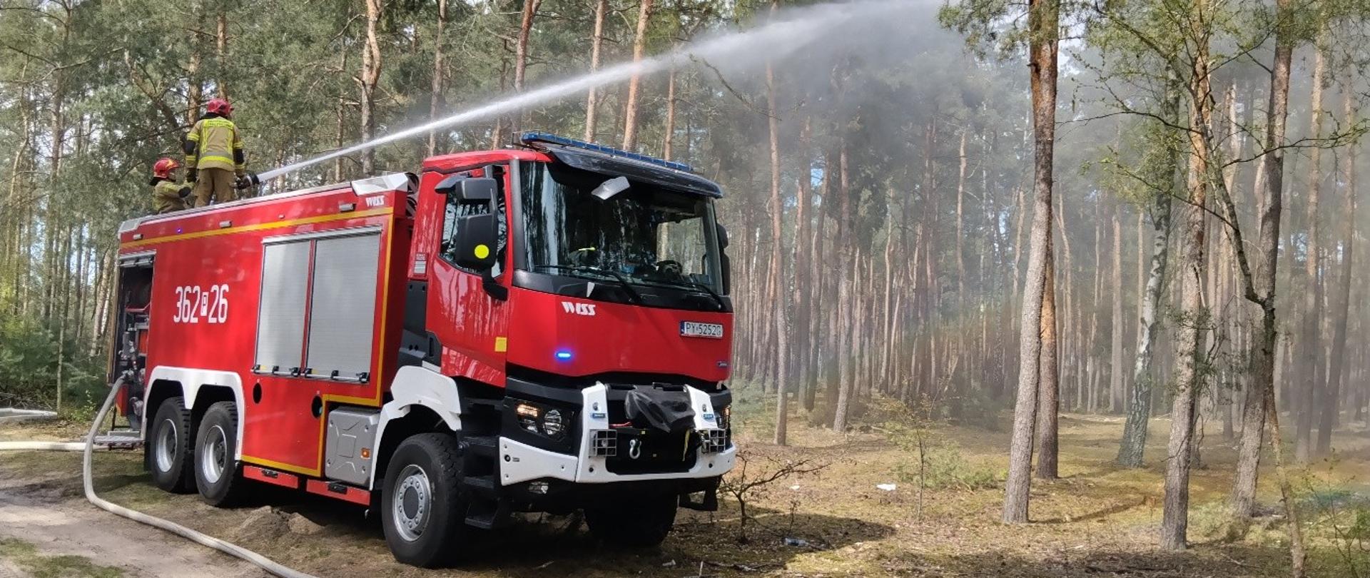 Strażak w umundurowaniu obsługuje autopompę samochodu pożarniczego. Samochód stoi na poboczu drogi leśnej. Podłączone są do niego węże ssawne i tłoczne.