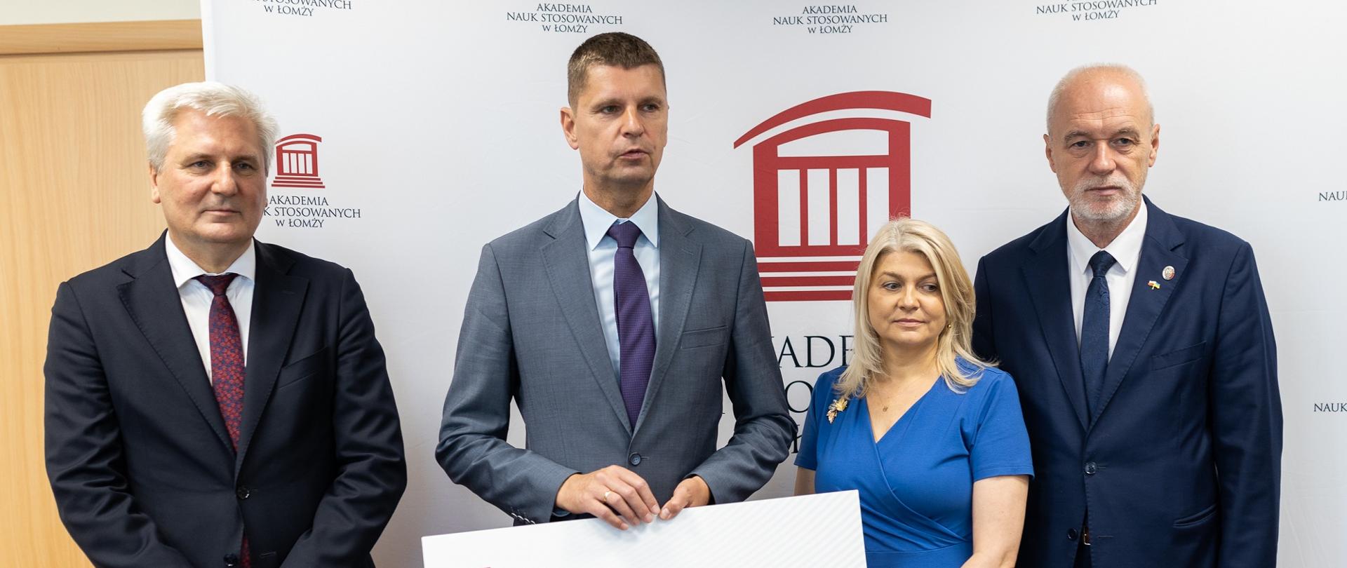 Minister Piontkowski stoi obok kobiety w niebieskiej sukni i dwóch mężczyzn w garniturach, trzyma symboliczny czek z napisem 4 436 900 zł.