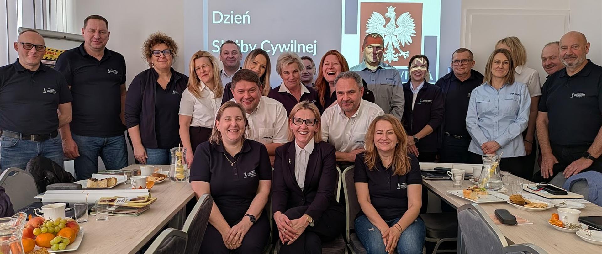 Grupa roześmianych osób pozuje do zdjęcia, w tle slajd 17 lutego dzień Służby Cywilnej
