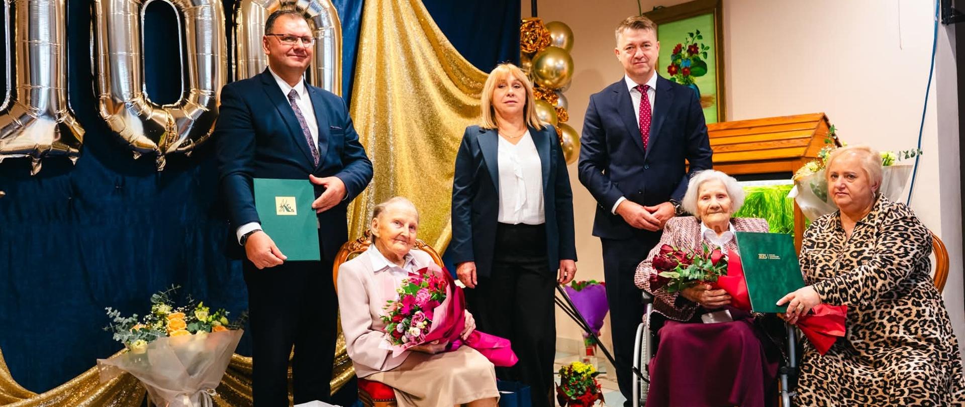 Grupa osób, trzy kobiety siedzą na fotelach, dwie trzymają bukiety kwiatów, za nimi stoi dwóch mężczyzn pomiędzy którymi stoi kobieta. W tle dekoracje, w tym srebrne balony - cyfry składające się na liczbę sto.