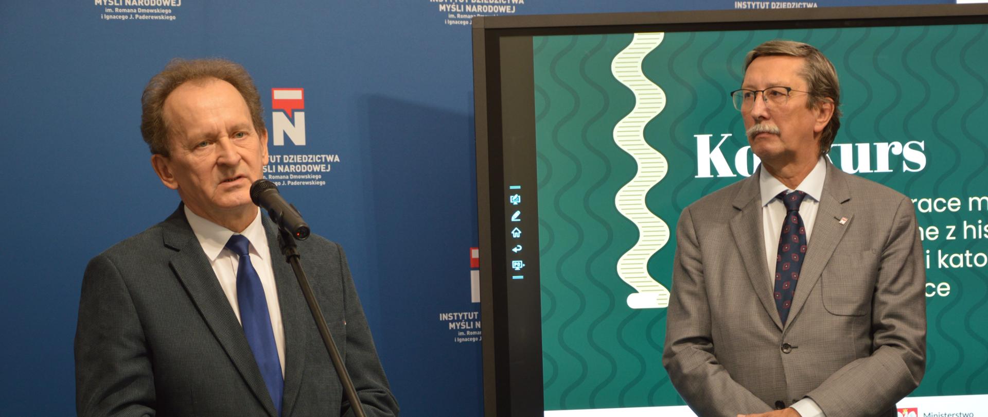 Wiceminister Bernacki mówi do mikrofonu. Obok niego stoi wiceminister prof. Żaryn za nim ekran z napisem Konkurs na najciekawsze prace magisterskie i doktorskie związane z historią ruchu narodowego i katolicyzmu społecznego w Polsce. W tle ścianka z logotypami Instytutu Dziedzictwa Myśli Narodowej im. Romana Dmowskiego i Ignacego J. Paderewskiego.