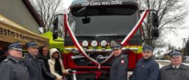 Grupa 7 osób pozuje na tle przodu samochodu strażackiego przepasanego biało-czerwoną wstęgą i napisem OSP KSRG Wielgorz. 