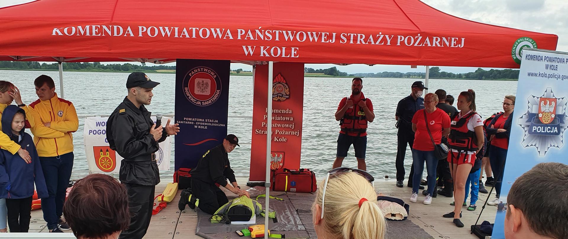 Zdjęcie przedstawia strażaków Państwowej Straży Pożarnej udzielających instruktażu pierwszej pomocy przedmedycznej oraz pokazu prowadzenia resuscytacji krążeniowo-oddechowej u dorosłego z użyciem automatycznego defibrylatora zewnętrznego „AED”. 