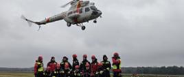 Grupa kilkunastu strażaków w ubraniach specjalnych i kaskach stoi i klęczy na płycie lotniska, a nad nimi na niewielkiej wysokości przelatuje śmigłowiec Straży Granicznej W-3.