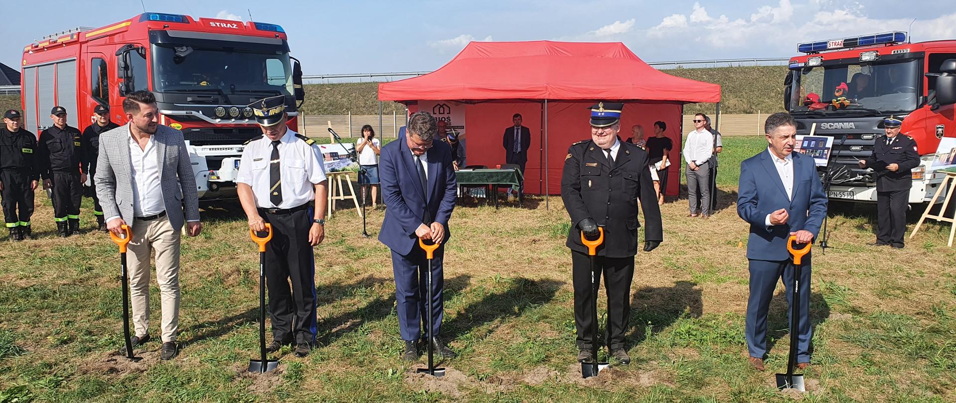 Pięciu mężczyzn, dwóch w ubraniach strażackich wyjściowych, trzech w garniturach, stoi za pięcioma szpadlami wbitymi w ziemię. W tle dwa samochody pożarnicze oraz czerwony namiot.