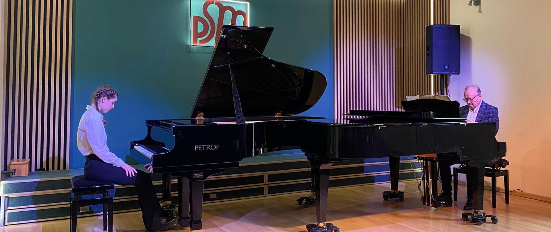 Zdjęcie przedstawia uczennicę siedzącą przy fortepianie po lewej stronie oraz pana dyrektora Artura Jaronia siedzącego przy drugim fortepianie po prawej stronie sceny, w tle widoczne zielona ściana z drewnianymi lamelami oraz logo szkoły