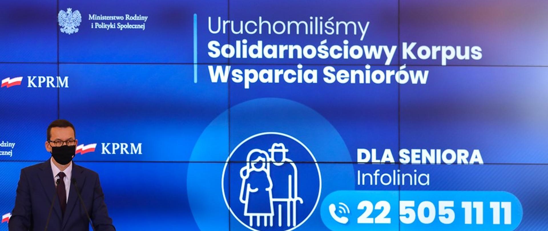 Premier Mateusz Morawiecki w ciemnym garniturze i w maseczce zakrywającej usta i nos podczas konferencji prasowej. Z tyłu niebieskie tło z napisem Uruchomiliśmy Solidarnościowy Korpus Wsparcia Seniorów. Infolinia dla seniora 22 505 11 11.