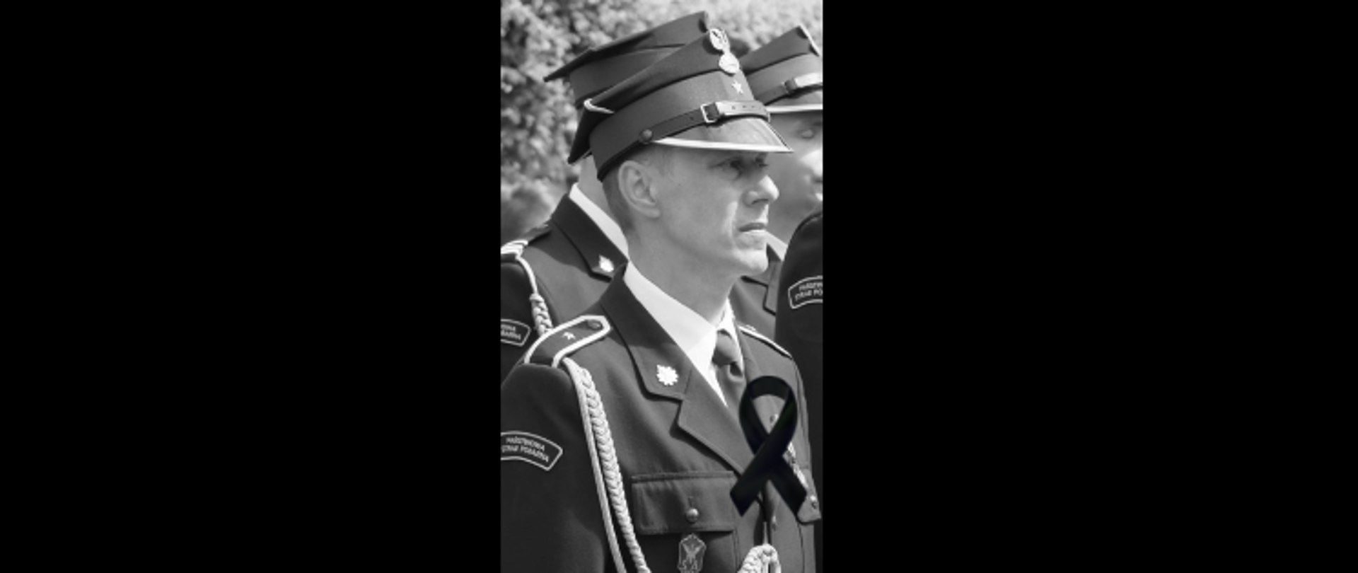 Strażak Państwowej Straży Pożarnej w galowym mundurze stoi w szeregu podczas uroczystości; czarno-białe ujęcie wykorzystywane w kontekście upamiętnienia zmarłego strażaka.