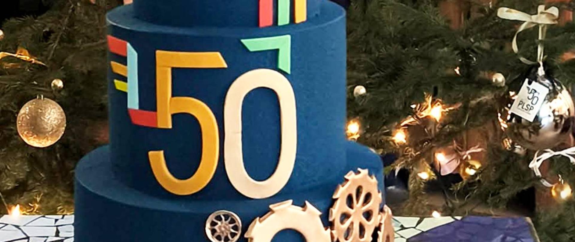 Cyfry 50 widoczne na niebieskim torcie, w tle fragment zielonej choinki i dekoracji świątecznych.